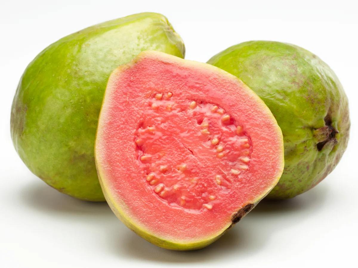 Due frutti di guava interi con buccia verde e una metà tagliata che mostra la polpa rosa brillante e i semi, isolati su sfondo bianco.